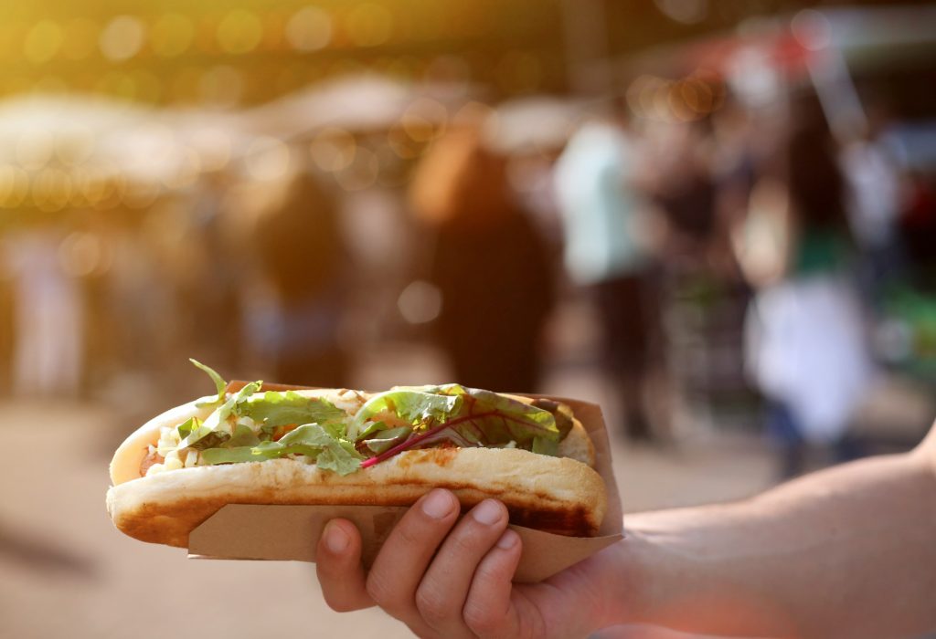 Close up of a hot dog in hand against blurred summer street food festival.